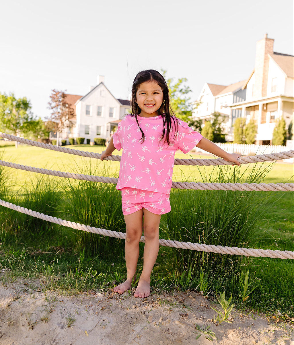 Young girl in a pink outfit standing on a sandy patch with a rope fence and houses in the background.