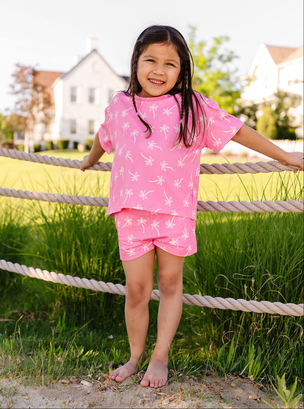 Young girl in a pink outfit standing outdoors near a rope fence with houses in the background.
