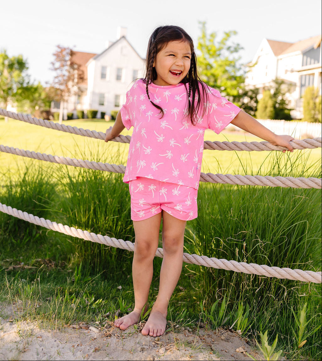 Young girl in a pink outfit standing outdoors near a rope fence with houses in the background.