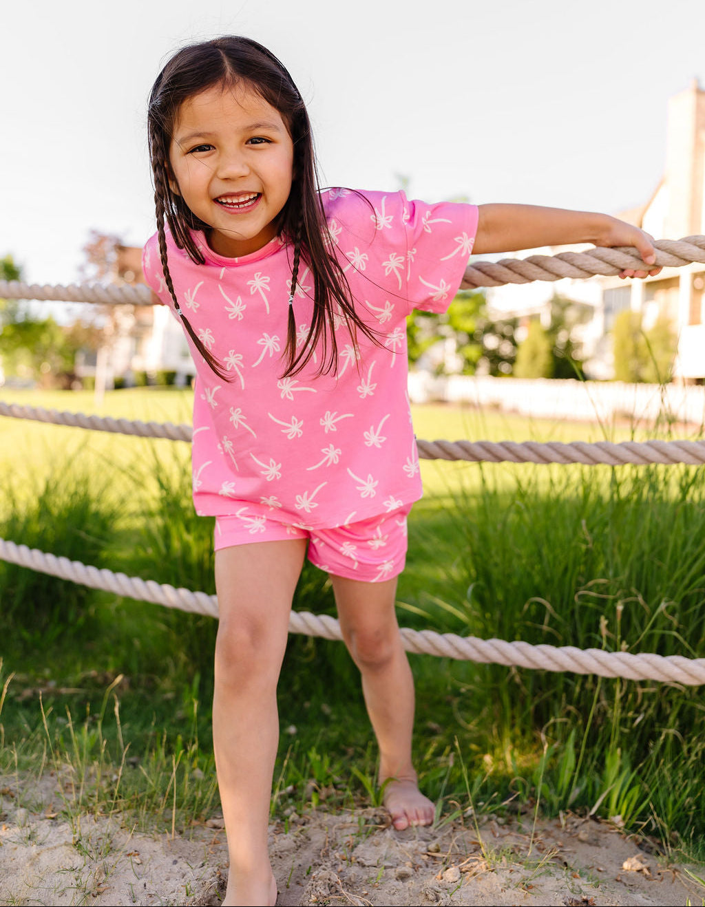 Young girl in a pink outfit standing on a sandy area with a rope fence and grass in the background.